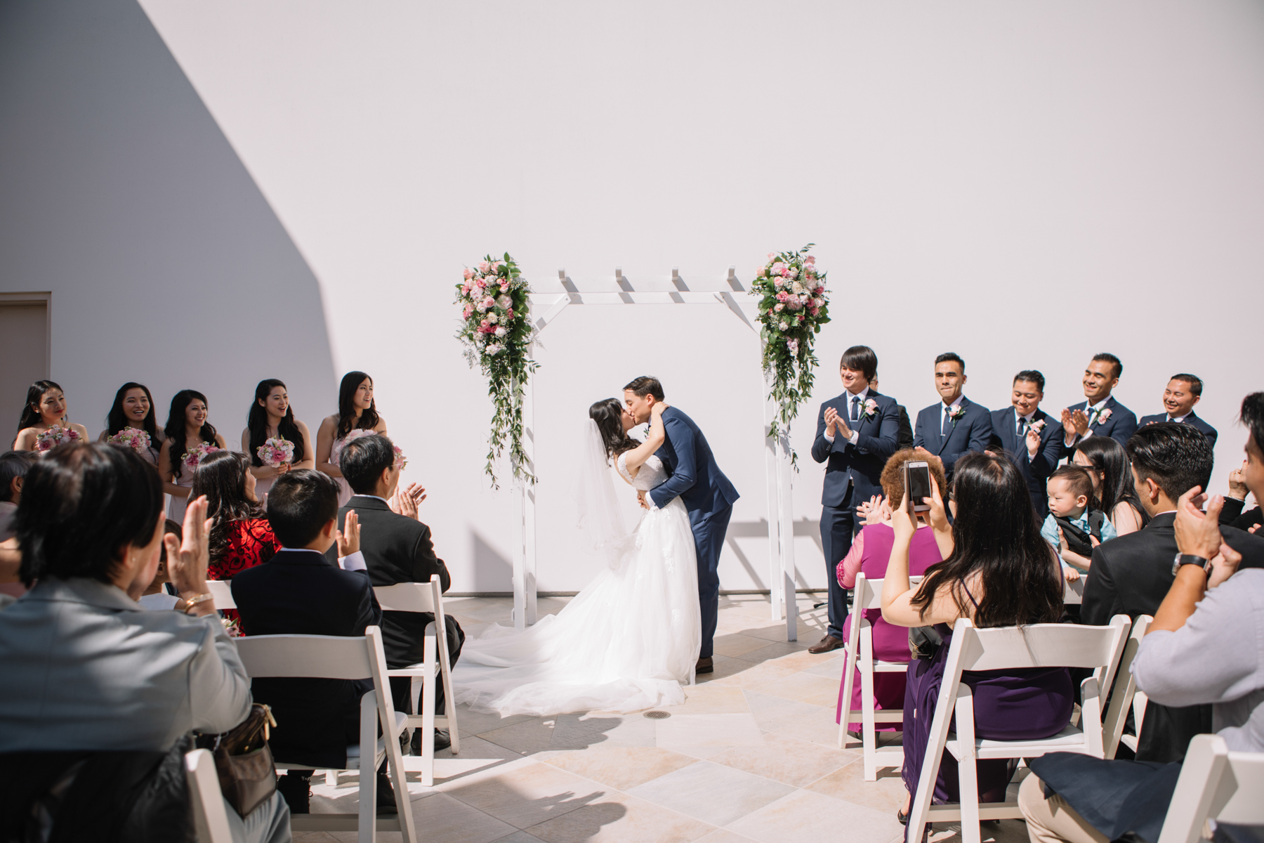 A small wedding venue outside with a bride and groom kissing in front of a white wall as their guests celebrate