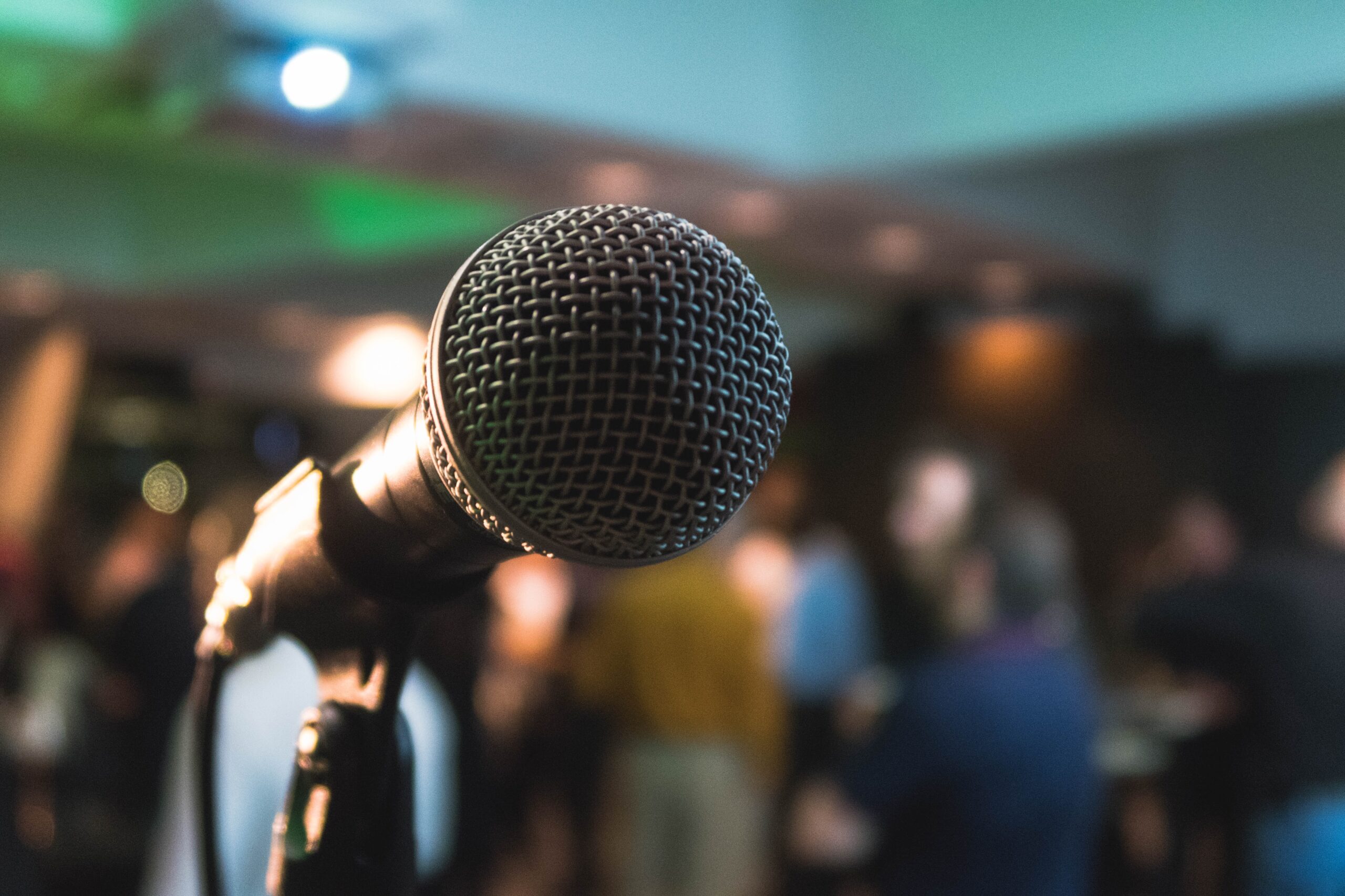 A close-up of a microphone with a business conference in the background filled with people listening. It is a perspective from stage out to the audience.