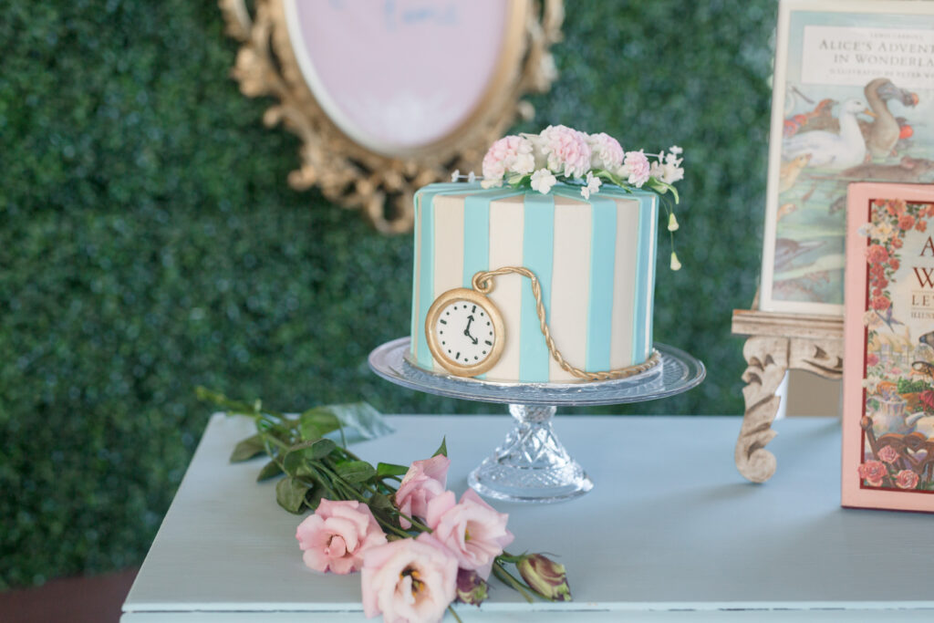 A cake decorated with blue and soft pink stripes with the watch indicating the Alice in Wonderland theme. 