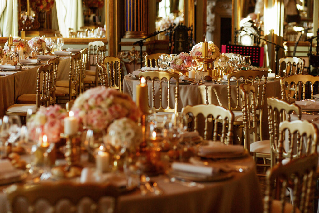 A gold event theme setup for an anniversary celebration in a beautiful ballroom with gold place settings and rose and gold flowers throughout