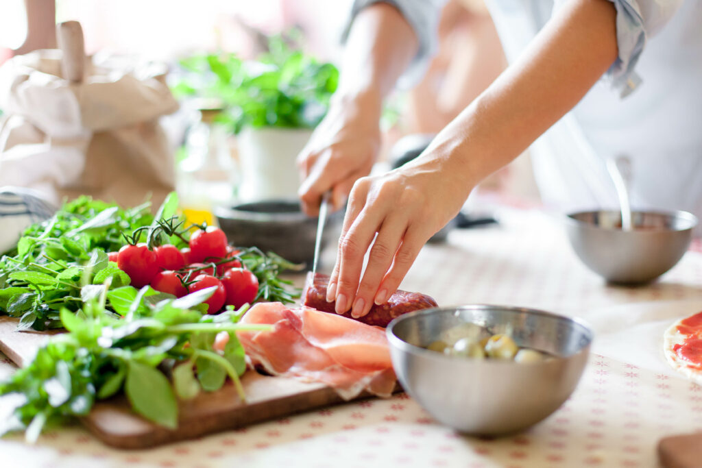 Woman is doing in a cooking demonstration. Female hands cut salami, vegetables, greens, tomatoes on table on wooden boards. Ingredients for preparing italian or french food. Lifestyle moment.