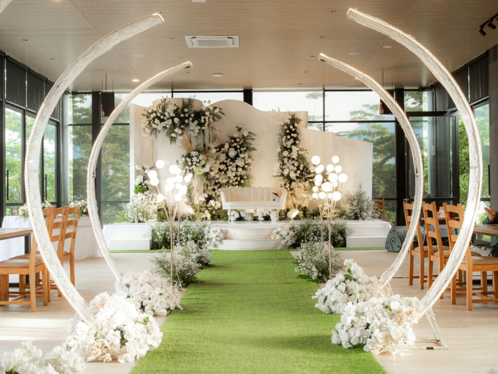 An indoor wedding with a beautiful aisle lined with a green flooring, similar to astro turf. There are also white flowers lining the aisle with beautiful 1/3 circles going over the aisle like an archway.